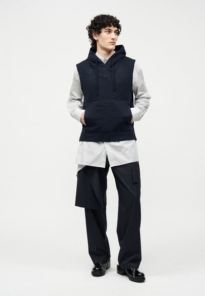 Jonge man met krullend haar, gekleed in een zwart mouwloos vest met capuchon over een wit lang shirt en zwarte wijd uitlopende broek met gelaagd stofdetail.