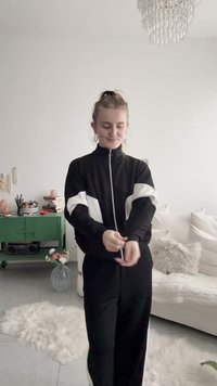 Jeune femme en survêtement noir et blanc ajustant ses manches dans un salon lumineux et chaleureux avec un canapé blanc, une armoire verte et un lustre.