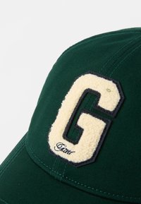 Cappellino da baseball verde con un'eccezionale patch in crema a forma di lettera "G" contornata in nero, con dettagli di cucitura visibili sul corpo.
