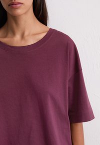 Close-up van een persoon in een losse, kortaf shirt in donkerpaars tegen een witte gestructureerde mu backdrop.