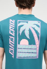 Mann trägt ein türkisfarbenes T-Shirt mit einem pink-blauen Motiv aus Palmen, Sonnenuntergang, Wellen und senkrechtem „Santa Cruz“-Schriftzug auf dem Rücken.