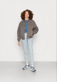 Casaco bomber cinzento com punhos canelados, t-shirt azul-claro, jeans de lavagem clara e ténis cinzentos com detalhes em preto. Estilo casual.
