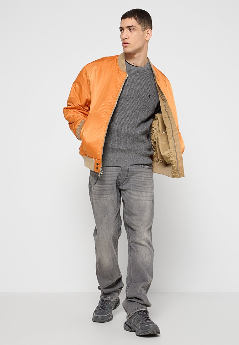 Junger Mann in einer orangefarbenen Bomberjacke über einem grauen Pullover, grauen Jeans und grauen Sneakers, der steht und zur Seite schaut.