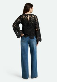 Top in pizzo nero con maniche a campana lunghe e orlo scallopato, caratterizzato da uno scollo a cuore sul retro. Abbinato a jeans in denim blu a gamba larga.