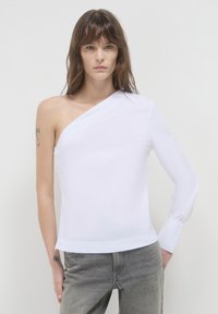 Frau trägt ein asymmetrisches Top mit einem weißen Ärmel und graue Jeans und steht vor einem schlichten hellen Hintergrund.
