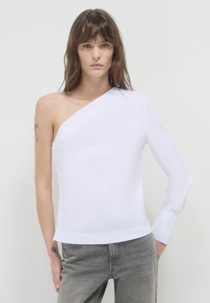 Frau trägt ein asymmetrisches Top mit einem weißen Ärmel und graue Jeans und steht vor einem schlichten hellen Hintergrund.