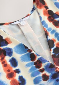 Večbarvna bluza z V-izrezom, narejena iz lahkega blaga z modrim, rdečim in črnim vzorcem na krem ozadju.