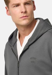 Sudadera gris con cremallera de textura suave, que cuenta con una capucha ajustada y un pequeño logo bordado. Llevada sobre una camisa blanca con cuello.