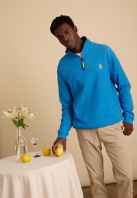 Blauwe fleece pullover met een halflange rits kraag, met een embleemlogo. Model houdt een citroen vast naast een tafel met een wit tafelkleed en bloemen.