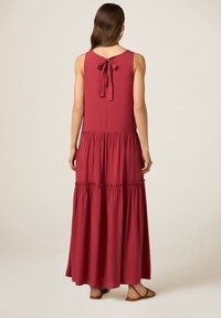 Abito lungo senza maniche in tessuto bordeaux. Presenta una chiusura con fiocco annodato sul retro, un design a strati e una silhouette fluida.