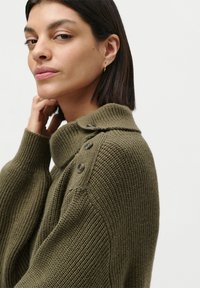 Frau mit kurzem dunklem Haar, die einen grünen Strickpullover mit Knöpfen an der Schulter trägt, den Hals berührt und leicht seitlich gegen einen einfarbigen Hintergrund schaut.