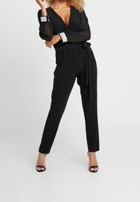 Kvinna som bär en svart jumpsuit med långa genomskinliga ärmar, djup V-ringning, paljetter i ärmsluten och ett knytbälte i midjan, i kombination med svarta öppna klackskor.