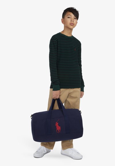 Bolsa de lona azul marino con logo rojo de jugador de polo, tela resistente, dos asas superiores y cierre con cremallera. El modelo lleva una camisa de manga larga a rayas verdes y pantalones beige.