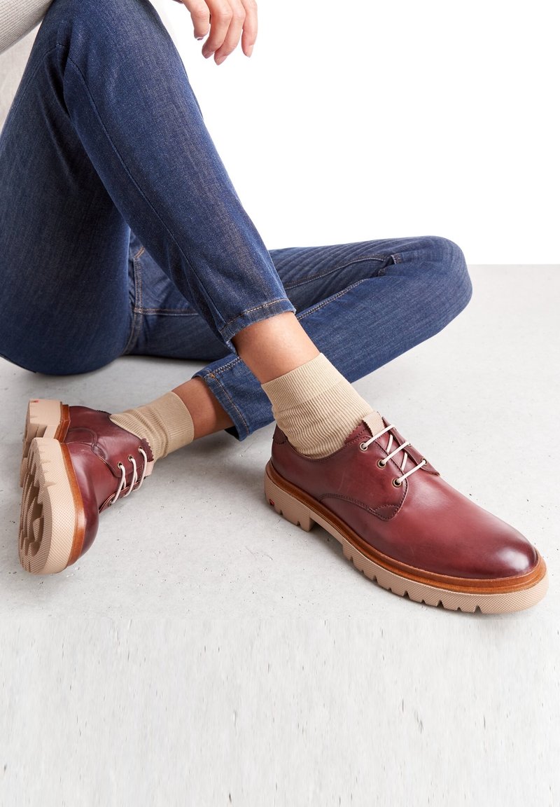 Chaussures en cuir rouge à lacets avec une semelle beige et un motif de semelle visible, associées à des chaussettes en coton côtelé beige et un jean bleu foncé.