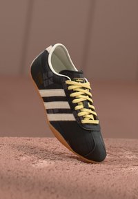 Black synthetic sneaker with a glossy finish, yellow laces, three white stripes, and a gum sole. Features a textured, patterned design.