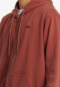 Hnedo-oranžová mikina na zips s predným vreckom, šnúrkami na kapucni a kontrastným logom na hrudi. Mäkkučký, štruktúrovaný materiál.