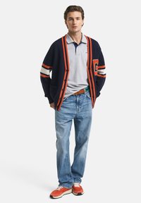 Námořnický svetr s oranžovými pruhy a detailním písmenem, šedý polo tričko, světle modré džíny a oranžové tenisky. Celkový casual design.
