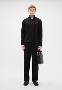 Sweater preto com fecho, calças largas pretas e sapatos pretos. Segurando uma mala de couro preta com um design estruturado. Marca mínima visível.