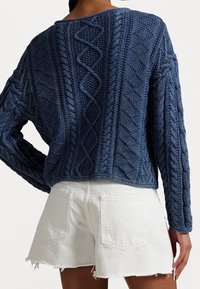 Blauer Pullover mit Zopfmuster, strukturiertem Design, kurz geschnitten und langen Ärmeln, kombiniert mit weißen, ausgefransten Shorts. Rückansicht gezeigt.