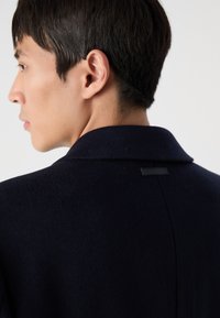 Marineblauwe wollen jas met een notch-kraag, met een gladde textuur en een klein zwart label op de achterkant. Gestructureerde pasvorm met strakke lijnen.