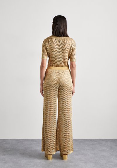 Combinaison en tricot doré avec un motif en chevron, dotée de manches courtes et de jambes larges. La taille est accentuée par une ceinture assortie.