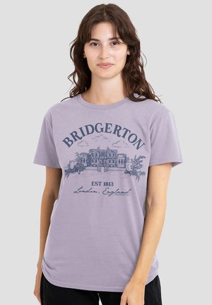 Young woman with long brown hair wearing a lavender Bridgerton T-shirt featuring a vintage mansion and text "EST 1813 London, England."
