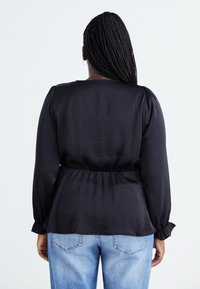 Blouse noire à manches longues avec une taille froncée et une coupe ample, dotée d'une texture douce et d'une légère brillance, associée à un jean bleu clair.