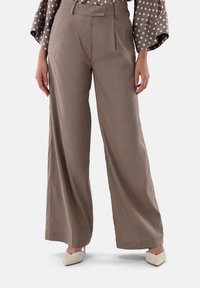 Hellbraune, weit geschnittene Hosen mit glatter Textur und gefaltetem Frontbereich. Kombiniert mit weißen High-Heels und einer gepunkteten Bluse.