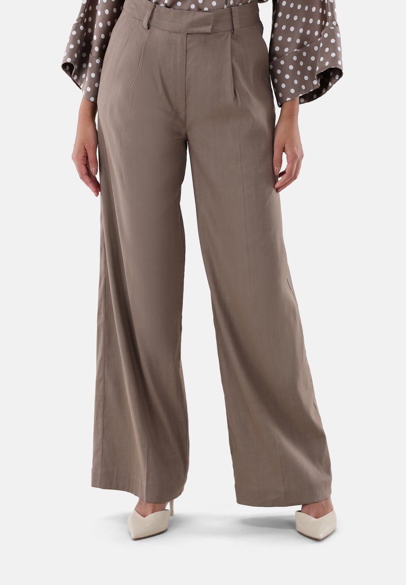 Hellbraune, weit geschnittene Hosen mit glatter Textur und gefaltetem Frontbereich. Kombiniert mit weißen High-Heels und einer gepunkteten Bluse.