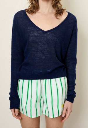 Femme portant un pull à col en V bleu marine à manches longues et un short blanc à rayures verticales vertes, debout devant un fond uni.