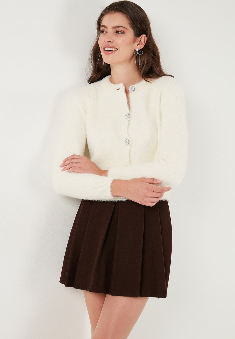 Bijela fluffy cardigan s okruglim izrezom, šest srebrnih dugmadi na zakopčavanju i dugim rukavima, u kombinaciji s smeškom naboranom mini suknjom.