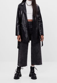 Czarny płaszcz z sztucznej skóry z klapami, noszony z białym topem crop, czarnymi spodniami z szerokimi nogawkami oraz czarnymi masywnymi butami sznurowanymi.