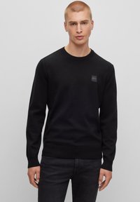Maglione nero a maglia con collo a girocollo e polsini a costine. Presenta un'etichetta con logo testurizzato sul petto. Abbinato a jeans scuri.