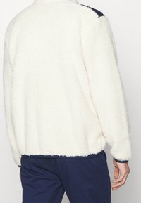 Fleecejacka i off-white, med en texturerad och mjuk yta samt ett marinblått accent vid axlarna. Dragkedja vid kragen och elastiska muddar.