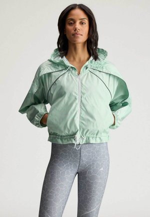 Mujer con chaqueta cortavientos con capucha de color verde claro y leggings gris con estampado floral, de pie con las manos en los bolsillos de la chaqueta.