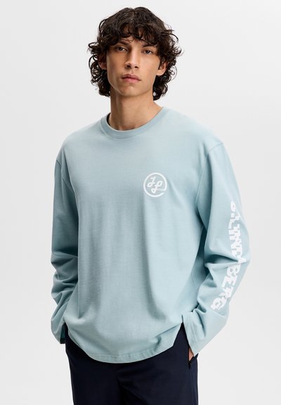 Lichtblauw T-shirt met lange mouwen gemaakt van katoen. Voorzien van een logo aan de voorkant en witte letters langs de linkermouw. Losse pasvorm met zijsplitsingen.