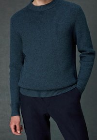 Ciemnoniebieski sweter z dzianiny o prążkowanej fakturze, z okrągłym dekoltem i długimi rękawami. W połączeniu z ciemnymi spodniami, podkreślającymi dopasowaną sylwetkę.