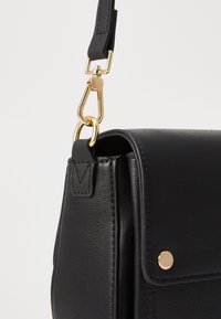 Borsa a tracolla nera in materiale sintetico testurizzato, con hardware dorato e una tracolla staccabile. Presenta una chiusura a patta e un dettaglio con bottone a scatto.
