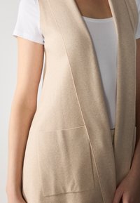 Beige stickad cardigan väst med öppen front, sidofickor och mjuk textur, bärs över en vit t-shirt. Har en ren, minimalistisk design.