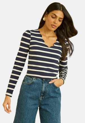 Navy and cream striped long-sleeve top with a scalloped neckline, worn with high-waisted blue denim jeans. Plain background.