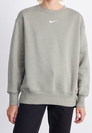 Personne portant un sweat-shirt Nike gris clair à manches longues et un pantalon noir, montré du cou aux hanches sur un fond uni.