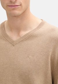 Suéter beige de cuello en V hecho de tejido de punto. Presenta un cuello acanalado y un pequeño logo bordado en el lado izquierdo del pecho. Textura suave.