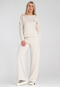 Jasno-beżowy zestaw odzieżowy składający się z topu z długim rękawem z kieszonką oraz spodni z szerokimi nogawkami, wykonany z miękkiego, teksturowanego materiału. Uzupełniony trampkami.