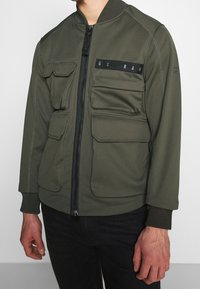 Khaki bomberjacka med flera framfickor, dragkedjestängning, ribbade muddar och en liten logotypapplikation på bröstet. Slät tygtextur.