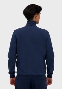 Sudadera azul marino con cuello alto, puños y dobladillo acanalados. Presenta un pequeño logo tricolor en la parte posterior del cuello. Tejido suave y texturizado.