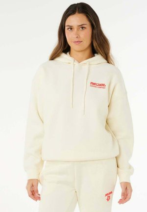 Frau trägt einen cremefarbenen Hoodie und passende Jogginghosen mit roten "Rip Curl Surfing Company"-Logos, steht vor einem schlichten Hintergrund.