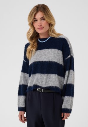 Femme aux cheveux blonds ondulés porte un pull duveteux à rayures bleu marine et gris, un pantalon foncé, une ceinture noire et un collier de perles bleu clair.
