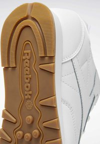Zapatilla deportiva blanca con suela de goma, que presenta un patrón de tread texturizado y la marca "Reebok". Parte superior suave con un sutil detalle de costura.