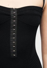 Κοντινό πλάνο μαύρου ριμπ πλεκτού τοπ χωρίς τιράντες με μεταλλικά κλείστρα μπροστά και ορατή τιράντα στον έναν ώμο.