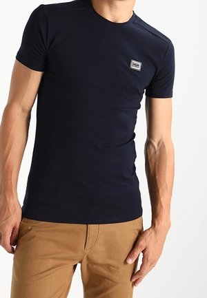 Mand iført en tætsiddende sort kortærmet t-shirt med et lille rektangulært logo på brystet og brune bukser mod en hvid baggrund.